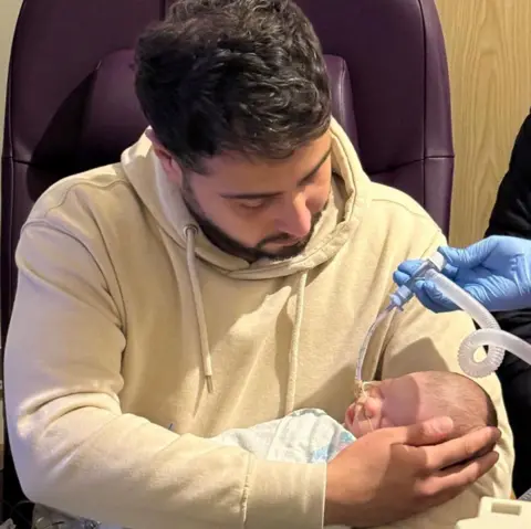 Hameed family A man with short black hair and beard is sitting in a chair as he holds a baby girl who has a breathing tube in her nose