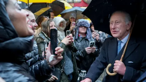 PA Media The King wears a suit and holds a black umbrella with a wooden handle. He is smiling as he greets shoppers. They are wearing winter coats, hats and some holding umbrellas as they smile in return and hold up mobile phones to document him, on a walkabout in Covent Garden.