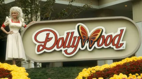 Ron Davis/Getty Images A smiling Dolly Parton poses in front of the Dollywood sign in 1988. She is wearing a long white dress with a red trim around the neckline and arms.