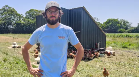 Chicken farmer Oliver Griggs, founder of Loma Farm in St John, Jersey stood wearing a t-shirt and cap outside one of his outdoor chicken pens. 