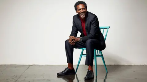 Hamish Brown Lemn Sissay, a man in a suit sitting on a green chair in an empty room, laughing. 