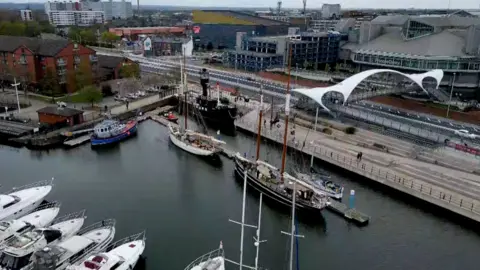 An aerial view shows a marina with white yachts and two tall ships moored at Hull Marina. Modern buildings, a road, and the white Murdoch's Connection bridge line the water’s edge in the background.
