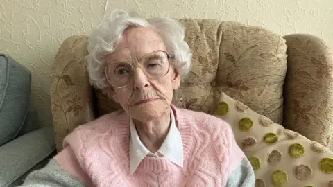 Photograph of Mary-Jane Conway, who survived a doodlebug attack in Tottington on Christmas Eve 1944. She is sitting in a chair at her care home. 