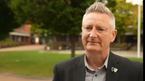 A man in a grey suit standing in front of trees outside Sandwell Council House.