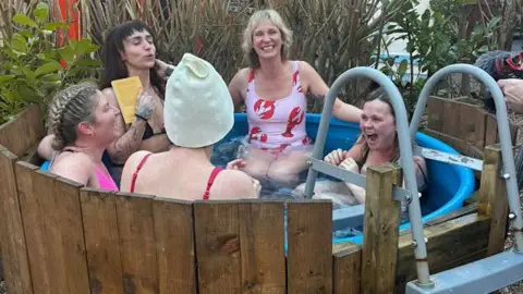Nicola Rees/BBC Five women wearing their swim suits sat in a round tub of water with wooden fencing round it. 