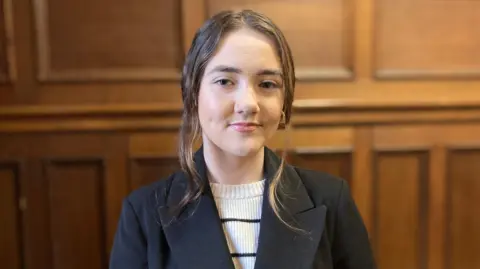 Dakota Nic Mheanman - a young woman with brown hair in an updo. She is wearing a black coat and a white and black stripped jumper. She is looking directly at the camera and is standing in front of a panelled wood wall.