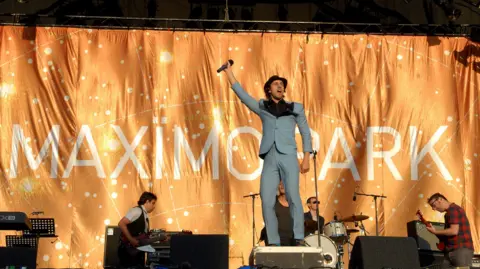 Maximo Park on stage in front of a large golden backdrop. Paul Smith looks to be standing on something in a bright blue suit and black hat. He has his hand in the air and is holding a microphone with his mouth open as if mid-song. The band is lower down around him and playing their instruments which include two guitars and some drums. 