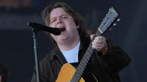 Lewis Capaldi sings into a black microphone while wielding brown six-string guitar. He is wearing a dark jacket and white t-shirt underneath.