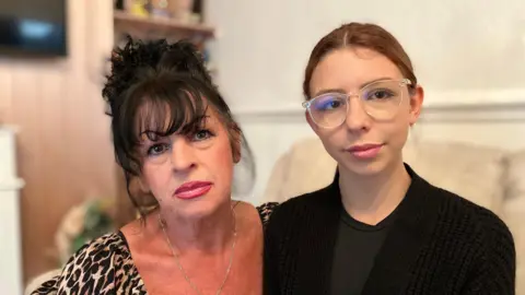 Sandra Bartlett has red lipstick and black hair. Deanna Ellis has light brown hair, big glasses and wears a black top and cardigan.