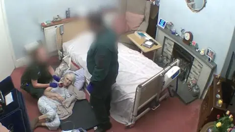 Kirstie Lumley  98-year-old women is lying on the floor having fallen out of bed.  She has two paramedics by her side looking after her.