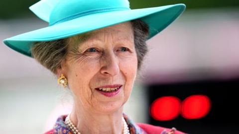 Princess Anne smiling, wearing gold earrings and a turquoise hat