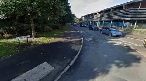 A Google street view image showing a road running between a building and a wooded area. Cars are queuing up on the right. A street sign on the left says Alexandra Road.