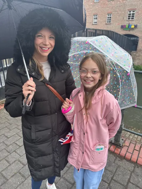 Joe Weir/BBC Sophia and her daughter Rose, standing under umbrellas in the rain. The woman is wearing a black coat with a hood, while the girl is wearing a pink jacket. Both are smiling.