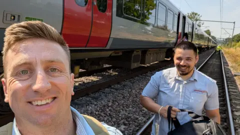 Jon Young Jon Young has taken a selfie with a member of Greater Anglia staff. Both are walking along the railway line next to the train and wearing pale blue shirts.