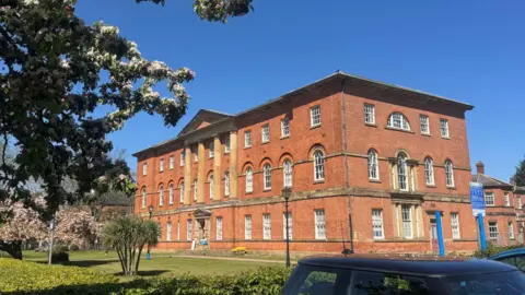 A large red‑brick building with tall front columns, arched middle‑floor windows and landscaped gardens with small palms and flowering trees under a bright blue sky.
