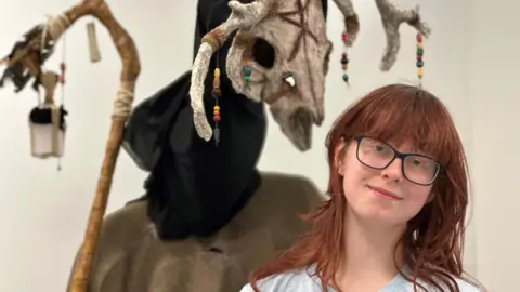 A mask with horns and a staff behind Charlotte Micklefield who has long red hair and glasses.