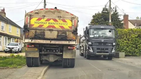 Two lorries try to pass each other on The Street in Cressing. The one on the left has a yellow and red high-visibility marking on its rear with the words "Highway Maintenance" written in bold type. It has mounted the kerb with two of its wheels. The lorry coming towards the camera is a black truck.