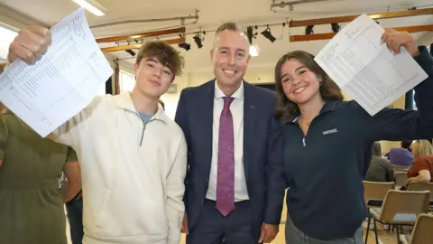 PA Media Paul Givan with a male pupil to the left and a female pupil to the right - both holding up their resuts