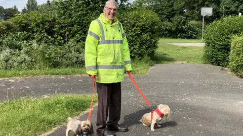 GUY CAMPBELL/BBC Dog owner wearing florescent yellow coat walking two small dogs on sturdy leads