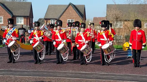 Soldiers in a red and black uniform play musical instruments including drums as they stand on the pavement outside the park.