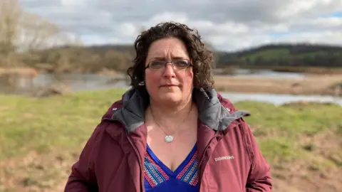 Gail Davies-Walsh looks at the camera as she stands in front of a green bank and water body. she is wearing a burgundy windbreaker coat, open in the front revealing a purple v neck blouse. she has rectangular glasses and ear length curly dark hair