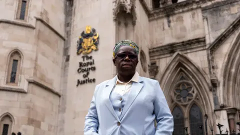 Jordan Pettitt/PA Media Rosamund Adoo-Kissi-Debrah in light blue suit stands in front of the Royal Courts of Justice. She wears a colourful headscarf, dark sunglasses. The building's stone façade and the words 