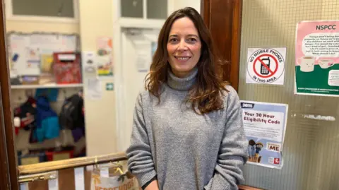 Chrissie Reidy/BBC Morgane Glew wears a grey polar neck sweater and stands in a nursery school smiling into the camera.
