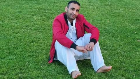 Image of Abdul Oryakhel. He is wearing white clothing, a red jacket and sitting on grass. He has short dark hair and facial hair and is looking directly at the camera and smiling.