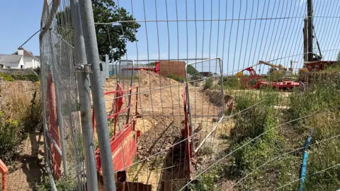 A picture o the scene where the sewage smell was reported. There is a large metal fence with grassland and building works in the distance.