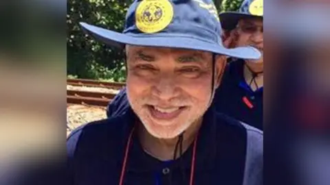 Supplied Dr Kumararatne is smiling at the camera while standing outside on a sunny day. He is wearing a navy hat and matching shirt.