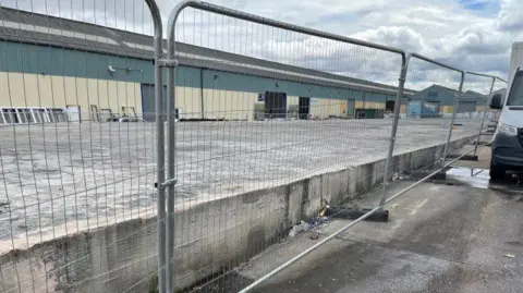 A fenced off section of an industrial estate. Just behind the fence is a concrete platform where the building that was lost to a fire once sat. Another factory building is visible in the background.