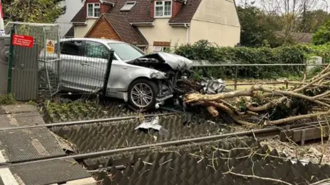 A white car has crashed through a fence and onto railway tracks with branches in front of it. The car's bonnet is badly damaged. 