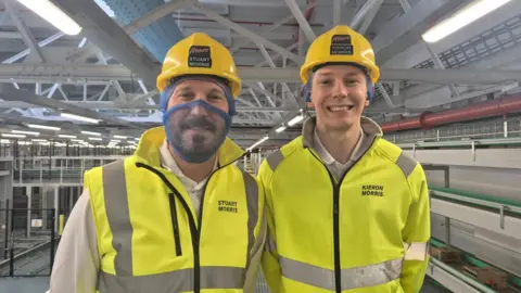 Father and son Stuart and Kieron standing side by side, facing the camera, smiling. Stuart is wearing a high visibility yellow gilet and is on the left. Kieron is on the right wearing a high visibility yellow jacket. Both are wearing white hard hats. Stuart has a blue food net over his nose and mouth