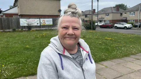 Tracey Walters smiling for the camera. There is a grassy area behind her with daffodils. She is wearing a nike jacket and a grey t-shirt underneath. She is wearing her hair as a bun on top of her head.