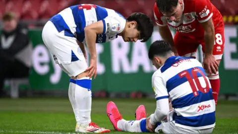 QPR's Koki Saito [left] and Middlesbrough's Aidan Morris [right] show concern for injured Hoops midfielder Ilias Chair [centre]