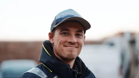 Matt Sharp Freddie Watson is a man with a blonde beard. He is smiling into the camera. He is wearing a blue cap and a navy waterproof with yellow trim. The background is blurred out behind him.