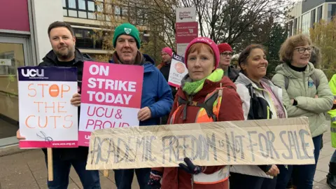 A group of men and women are holding placards and signs as they form a picket line outside Sheffield Hallam University