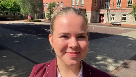A young girl is smiling toward the camera. She has blond hair tied up in a pony tail. She is wearing a maroon coloured school blazer and a red and white stripe shirt. Behind her is an open stone courtyard and a number of red brick buildings.  