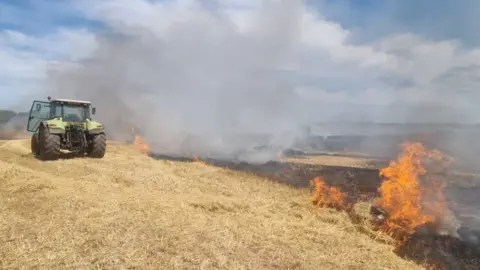 A field, covered in yellow, dried out grass, is partially on fire, with flames and smoke visible to the right of the image and in the background. In the upper left of the field, an abandoned green tractor can be seen