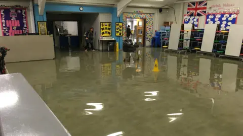 Russell Scott Primary School An assembly hall in Russell Scott Primary School filled with water and warning cones as teachers wade through the shallows. 