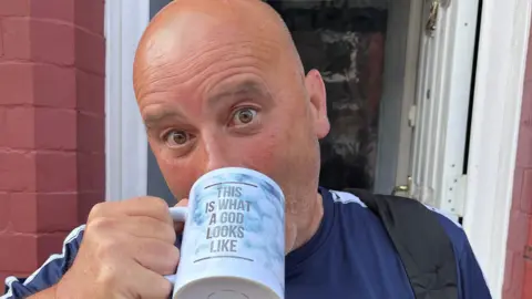 Family photograph The late Joseph Tidmarsh, who was bald and had a slight stubble. He is photographed wearing a navy and white t-shirt. He is standing outside an open front door while drinking out of a pale blue mug bearing the slogan: 'This is what a god looks like'.