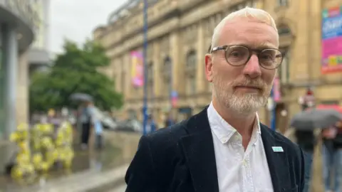 A close up of Peter Boulton. He has short blonde hair, is wearing glasses, and has a short beard. He is wearing a dark jacket and white shirt. In the background is the flower display.