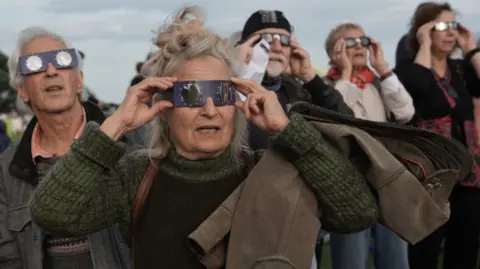 A group of older people look towards the sun in what appears to be mild amazement while clutching pairs of special glasses designed to protect their eyes from damage. The rims are made from card, with circular spectacles in the middle.