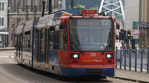 A red tram travels up hill. The driver can be seen through the glass of the cab