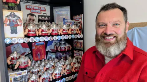 Jamie Moakes is wearing a red shirt and smiling. To the left is a cabinet full of Ram Man action figures, badges and posters. Ram Man is muscular and has silver shoulder plates, a red tunic and green trousers. 