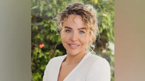 Lydia Bright wears a white top with lace trim. She has blonde curly hair which is tied back. She is sitting in front of a blurred background of green foliage.