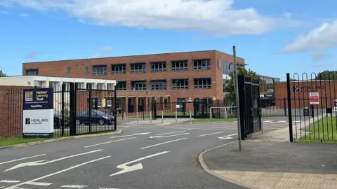 A red-brick, three-storey school building with black windows behind black gates. A grey road runs through the open gates. A sign reads: "Harbour Learning Trust, Healing Academy."