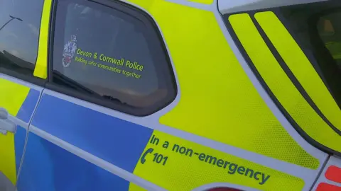 The rear end side of a police car with the Devon and Cornwall Police branding on the side. It has a non-emergency number on the side also. 