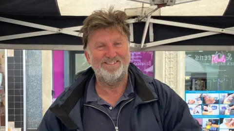 Henry Godfrey-Evans/ BBC A close-up of Trevor, who has light brown messy hair and a silver beard. He is wearing a navy coat and is standing under a gazebo.