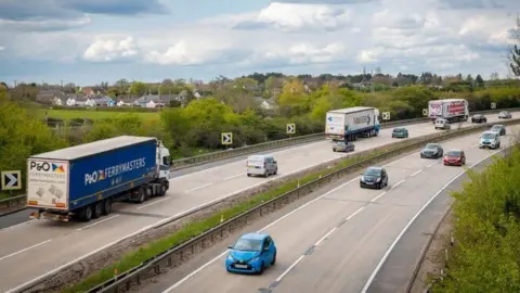 National Highways Cars and lorries drive on the A12 between Chelmsford and Brentwood. There are two lanes going in each direction. There are trees and bushes on both sides of the road and houses in the background.